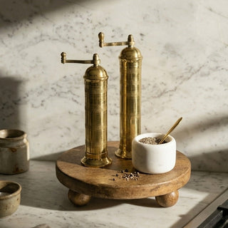 Two brass pepper grinders on a wooden stand with a marble background